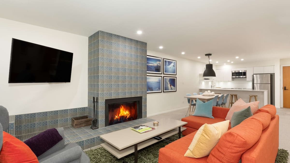 Living area with large orange couch with colorful cushions and large fireplaces with tiled pattern on it. White modern kitchen in background