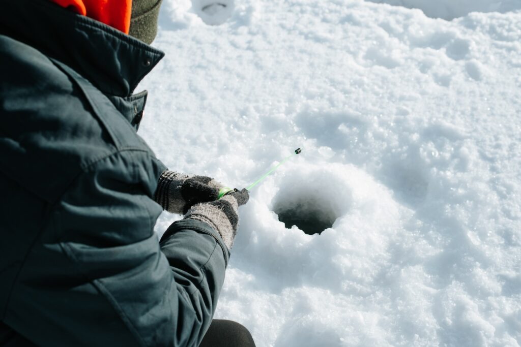 Winter fishing, active leisure, hobby and sport concept. Close-up of fisherman sitting on frozen water near drilled hole in ice. Selective focus on male hands in gloves holding fishing rod.
