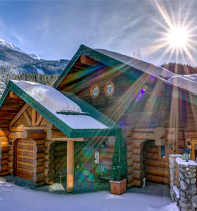 a bright sun shines behind a snow-covered log cabin in whistler, ca