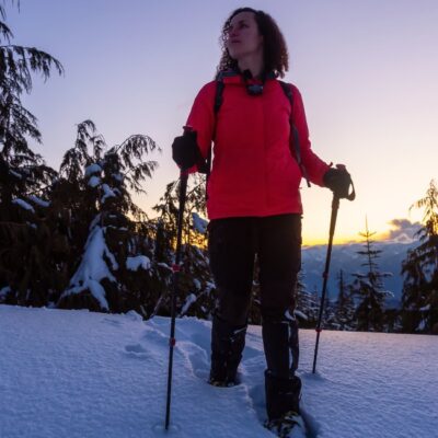 Adventurous woman snowshoeing in Whistler, with a picturesque view of snow-covered trees and distant mountain peaks illuminated by the setting sun.