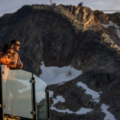A couple enjoying panoramic view from glass balcony at Whistler Blackcomb, overlooking snow-dusted Coast Mountains