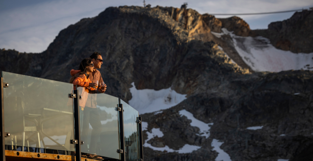 A couple enjoying panoramic view from glass balcony at Whistler Blackcomb, overlooking snow-dusted Coast Mountains