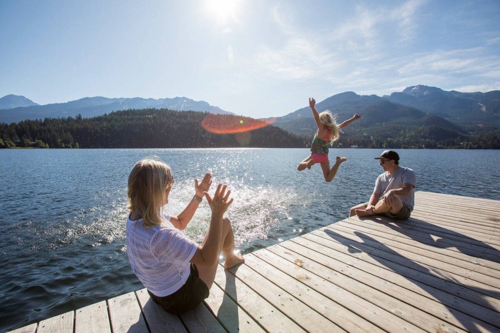 Family enjoying a sunny summer day at a lake in Whistler, BC—one child mid-jump into the water from a wooden dock, with others relaxing and soaking in the alpine scenery.
