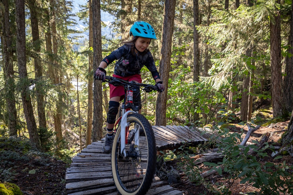 Young mountain biker navigating a narrow elevated wooden trail through lush forest in Whistler, BC, wearing a blue helmet and red-black gear, showcasing the area's world-class biking terrain.