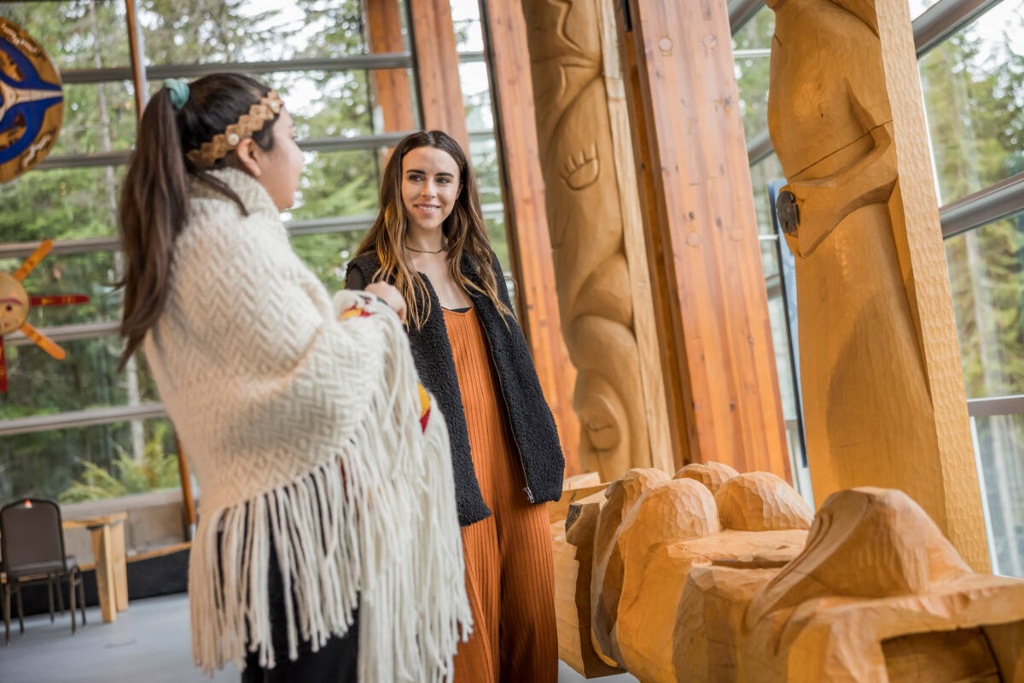 Visitors exploring an indoor exhibit of intricate wooden carvings at the Squamish Lil’wat Cultural Centre in Whistler, BC, with forest views through large windows and wooden architectural elements reflecting local Indigenous heritage.