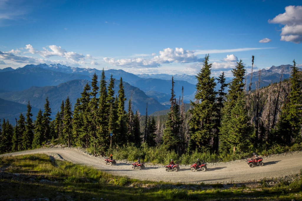 Five people riding red ATVs along a winding dirt trail through alpine forest, with panoramic mountain views and blue skies in Whistler during summer.